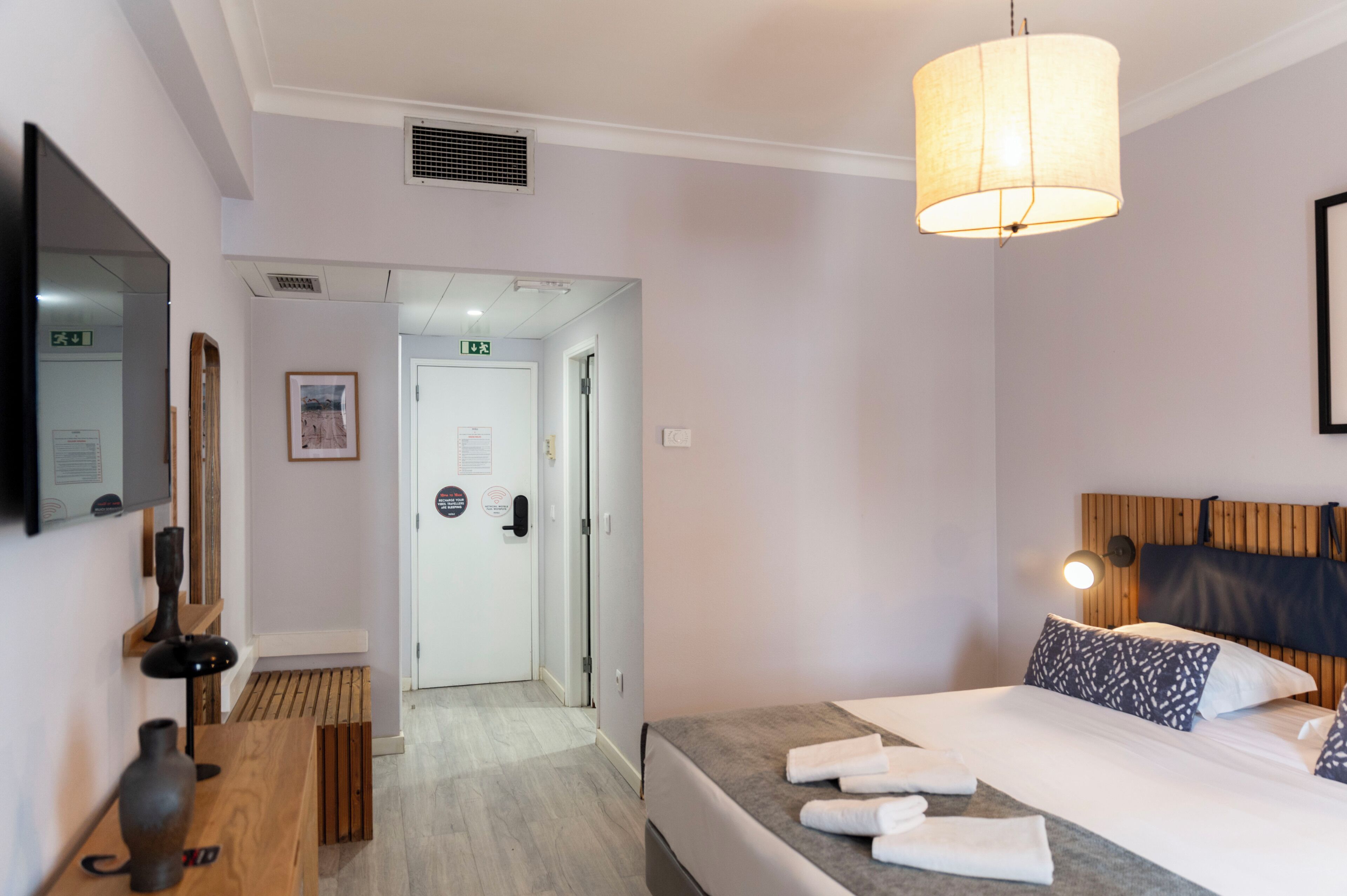 A hotel room with a bed, a TV, a wooden desk, a lamp, and a door leading to another room.