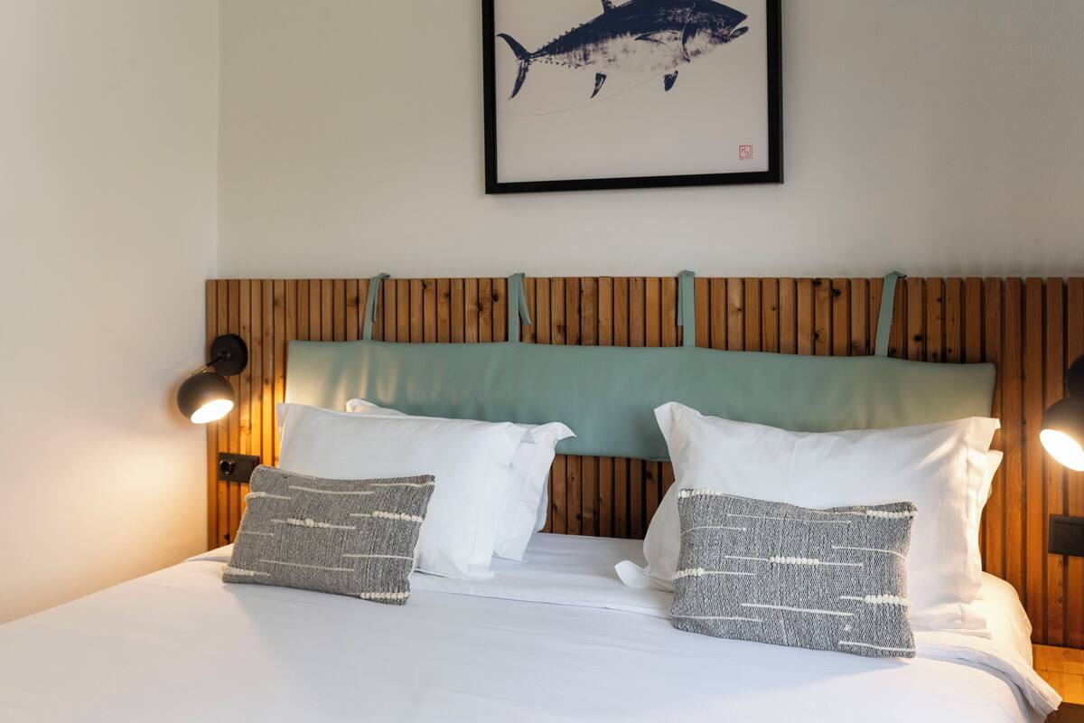 Un lit avec du linge de lit blanc, deux oreillers, une tête de lit en bois et une œuvre d’art représentant un requin, encadrée, accrochée au mur.