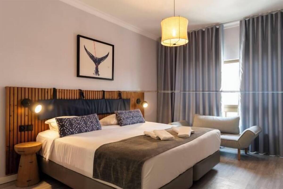 Une chambre d’hôtel moderne avec un grand lit, une tête de lit en bois, une table de chevet, une chaise et une grande fenêtre avec des rideaux.
