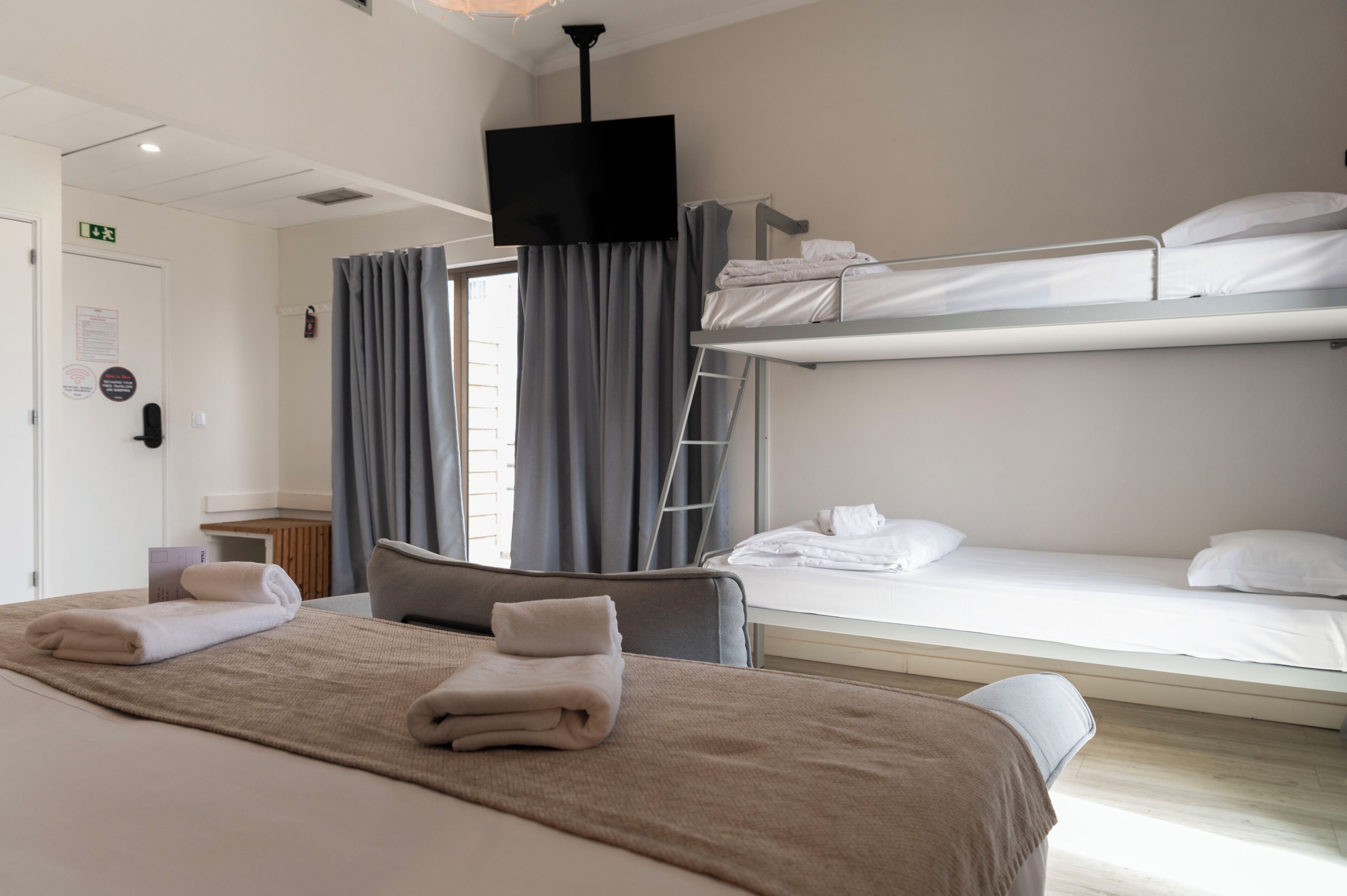 Une chambre d’hôtel avec un lit simple, un lit superposé, une télévision et une fenêtre avec des rideaux.