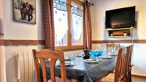 A dining area with a table set for four, wooden chairs, a television mounted on the wall, and a window with curtains.