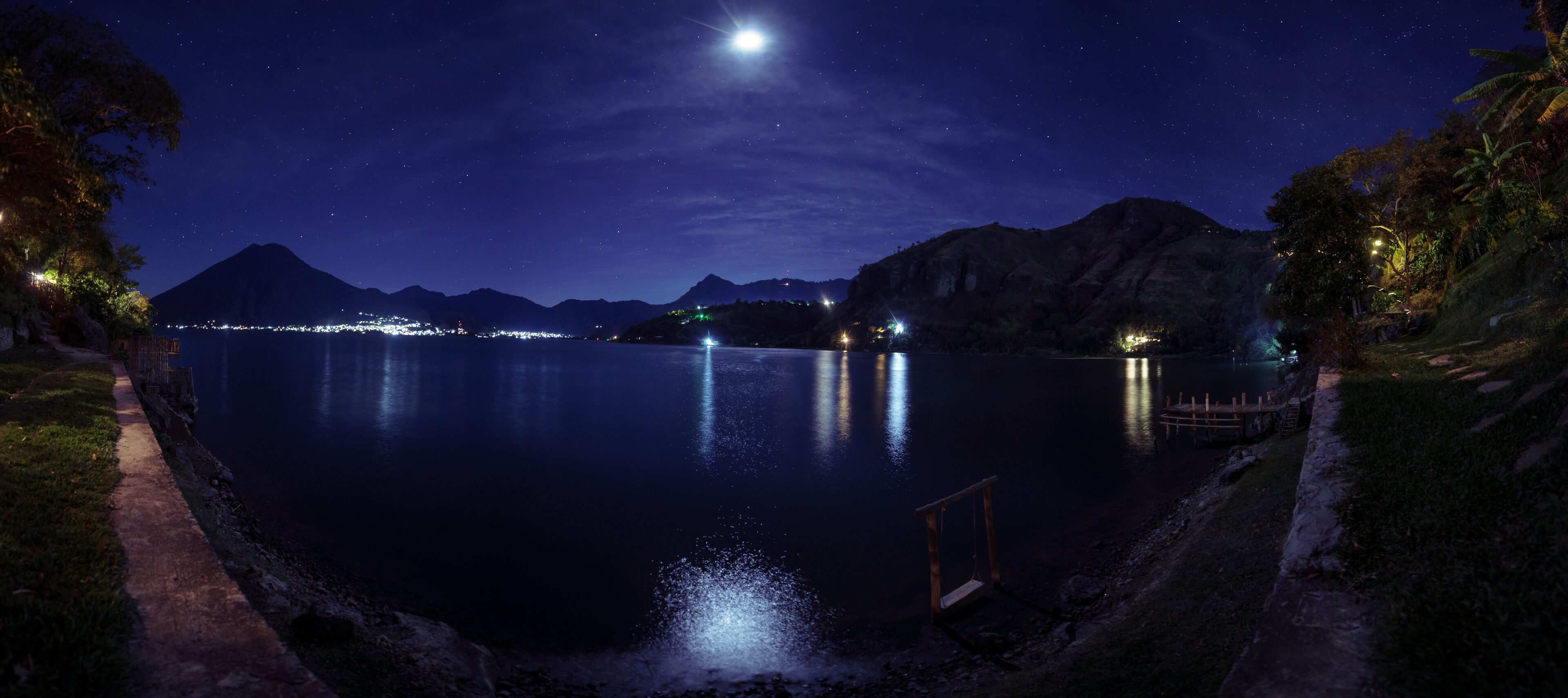 Nachtansicht eines Sees bei klarer Himmel, ruhiges Wasser spiegelt Lichter wider, und entfernte Berge sind zu sehen.