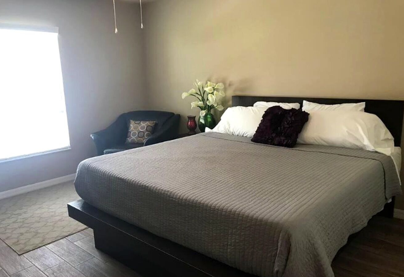 Une chambre à coucher avec un lit, une chaise, un vase de fleurs et une fenêtre.
