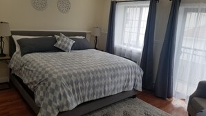 A bedroom with a large bed, two wall decor pieces, a window with curtains, and a nightstand with a lamp. - 20-2 Comfortable Quality Cribs (Queens)