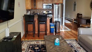 A modern kitchen with wooden cabinets, a stainless steel refrigerator, and a microwave. There are two bar stools, a trash can, and a table with a tissue box. - 20-2 Comfortable Quality Cribs (Queens)