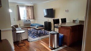 A compact living space with a kitchenette, a dining area with a table and chairs, a sofa, a TV mounted on the wall, and a trash bin. - 20-2 Comfortable Quality Cribs (Queens)