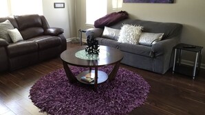 Una sala de estar con un sofá de cuero marrón, un sofá gris con cojines decorativos, una mesa de centro de vidrio y una alfombra morada.
