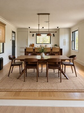 A modern dining area with a wooden table and chairs, a kitchen in the background, and large windows. - Bear Mountain House Near Hiking Trails (Philipstown)