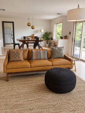 A modern living room with a sofa, ottoman, and a dining area in the background. - Bear Mountain House Near Hiking Trails (Philipstown)