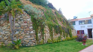 A stone retaining wall with vegetation, a grassy lawn, and a modern building with windows. - Complesso Il Saraceno (Maracalagonis)