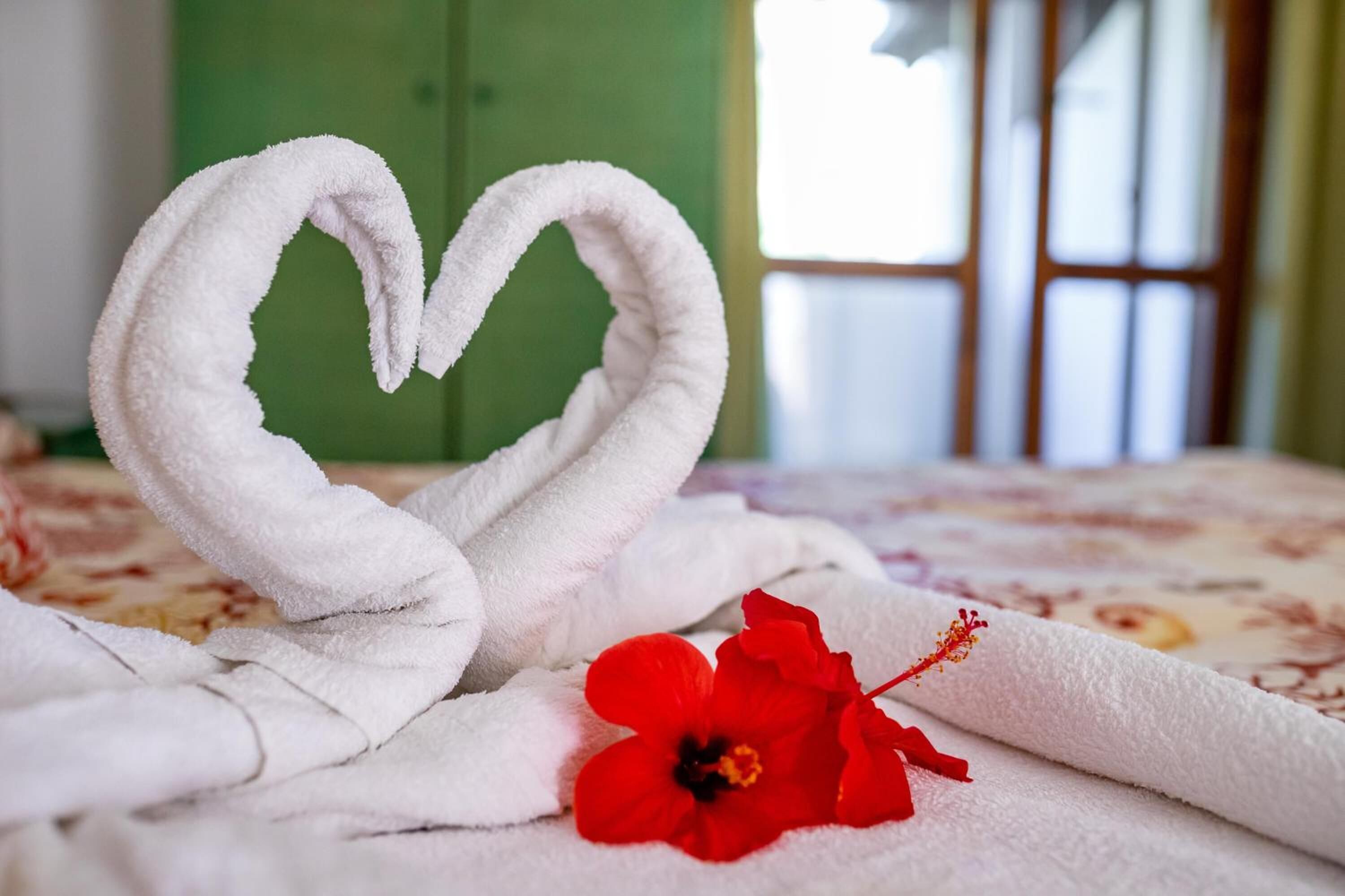 Una cama con una toalla dispuesta en forma de corazón y una flor de hibisco rojo.