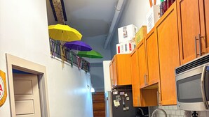 A narrow kitchen with wooden cabinets, a refrigerator, a microwave, and a sink. There is a chair and a table in the foreground. - OUR 1B/1B is PERFECT for your NOLA STAY! PARKING Avail! (New Orleans)