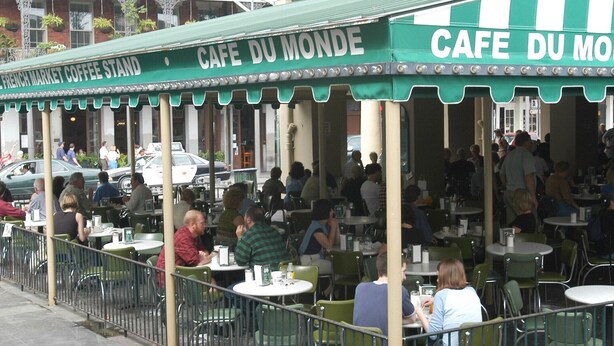 An outdoor café with tables and chairs, people dining, and a green awning with text. - OUR 1B/1B is PERFECT for your NOLA STAY! PARKING Avail! (New Orleans)