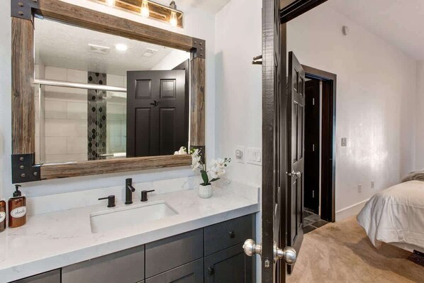 A bathroom with a large mirror, dark cabinets, a white sink, and a potted plant.