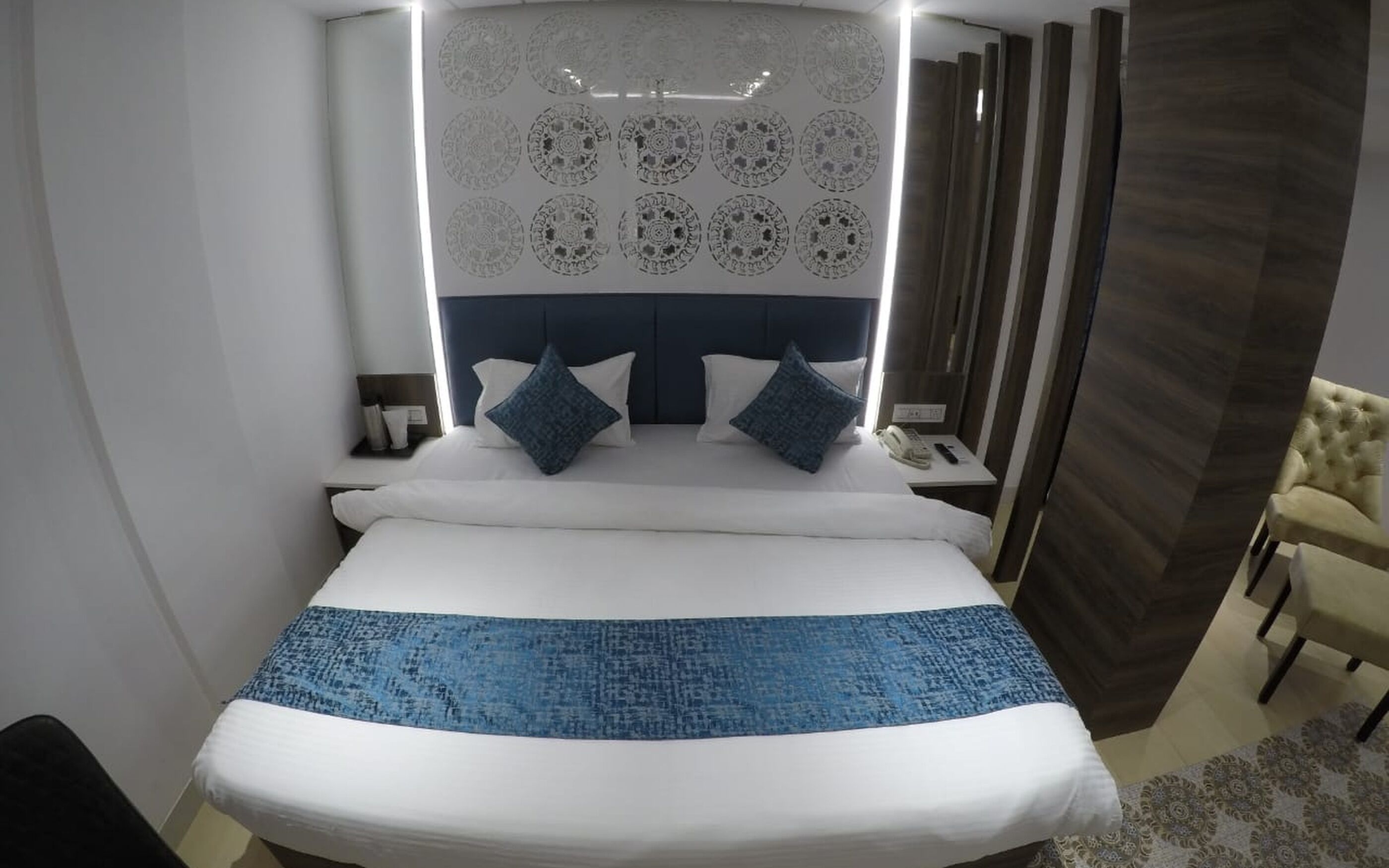 A modern hotel room with a large bed, a headboard with decorative patterns, a bedside table with a lamp, and a chair.