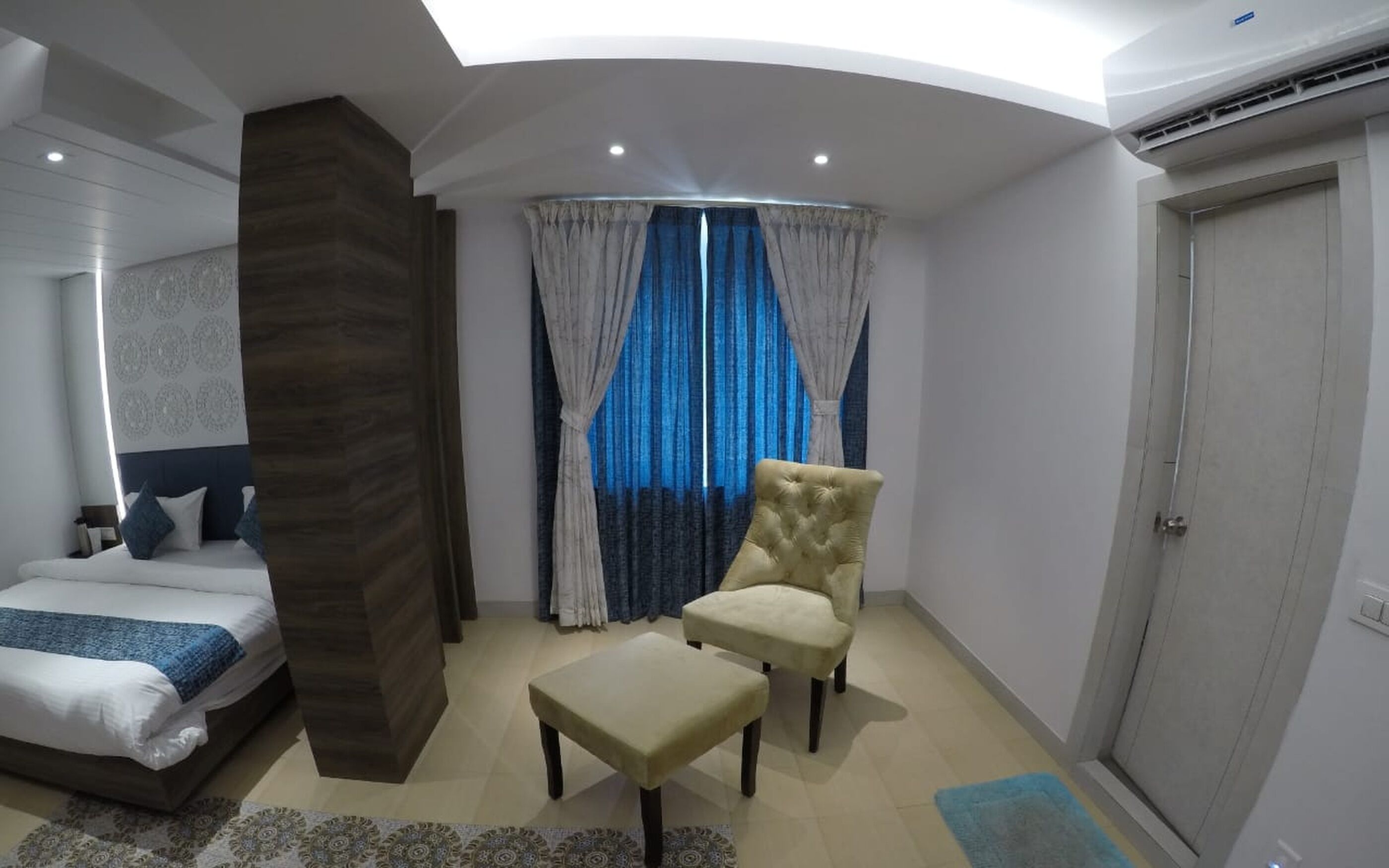 A modern hotel room with a bed, a large pillar, a chair, and a small table.