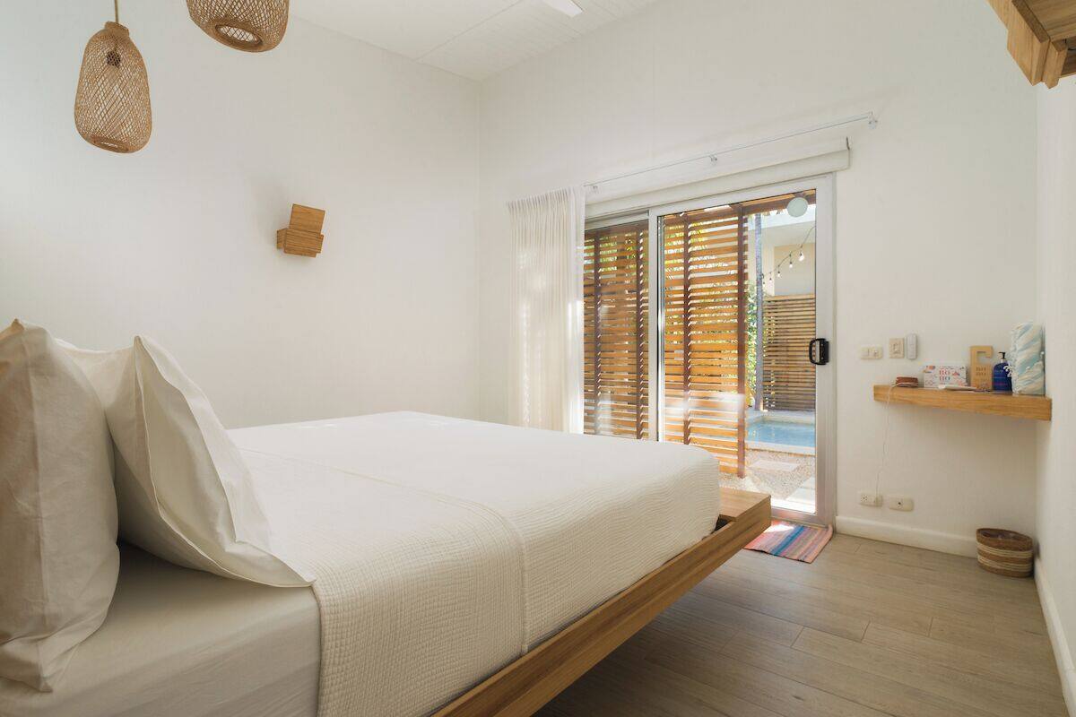 Ein Schlafzimmer mit Bett, Holzboden, einem Fenster mit Jalousien, einer zur Außenseite führenden Tür und einem Regal mit Toilettenartikeln.