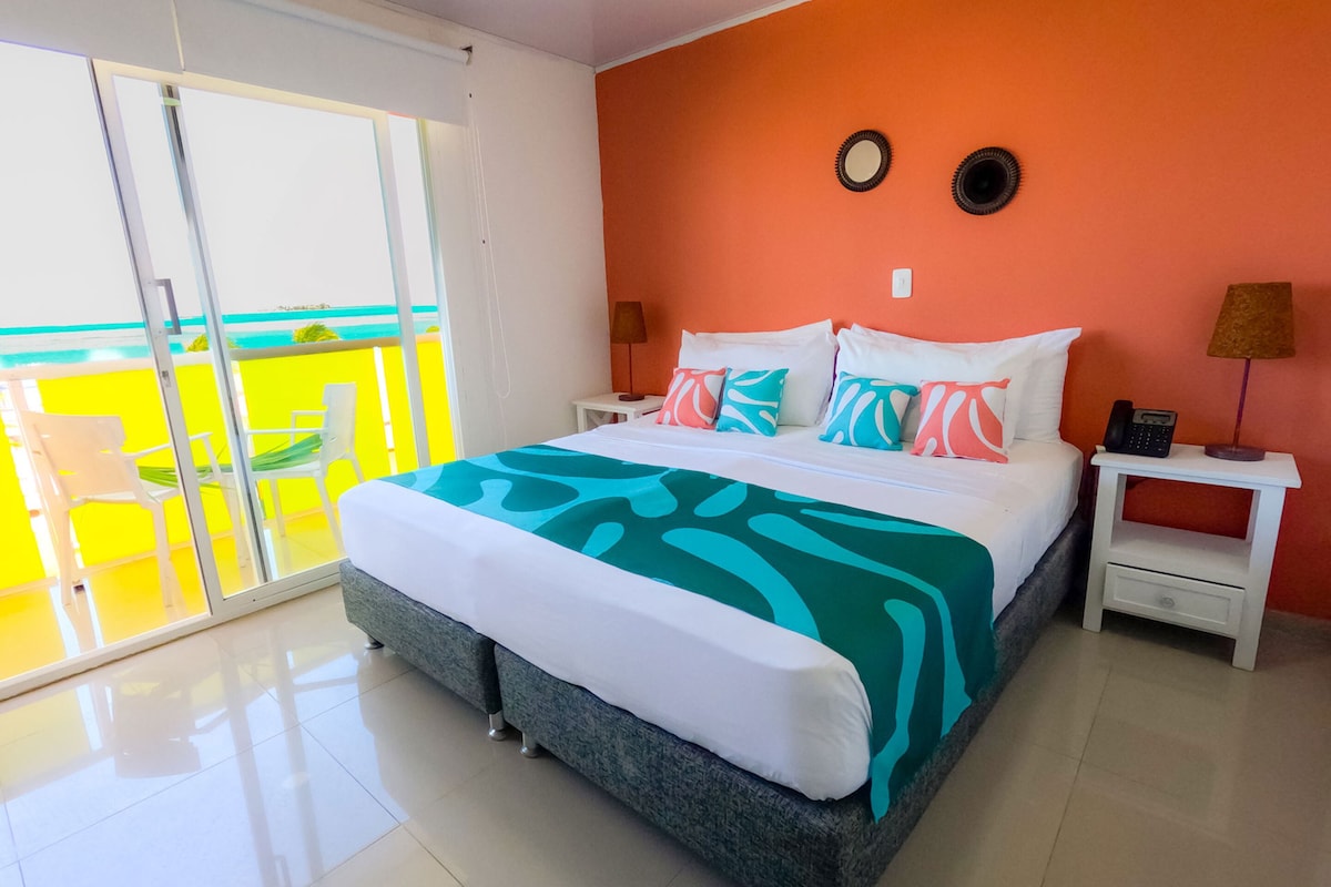 A bedroom with a bed, a nightstand, a lamp, a small table, and a view of a balcony with chairs.