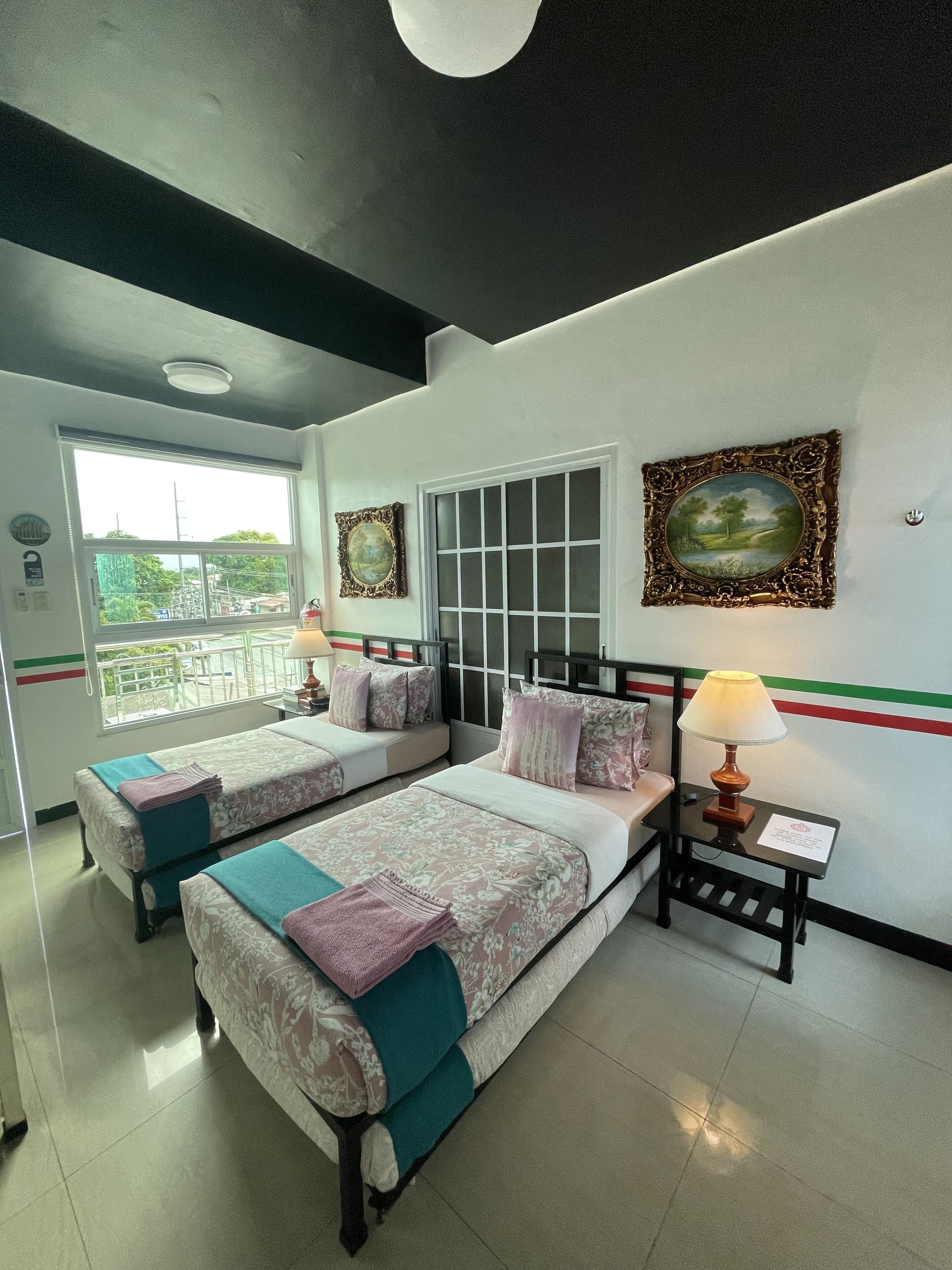 Habitación de hotel con dos camas, una ventana con vistas, una lámpara y dos cuadros en la pared.