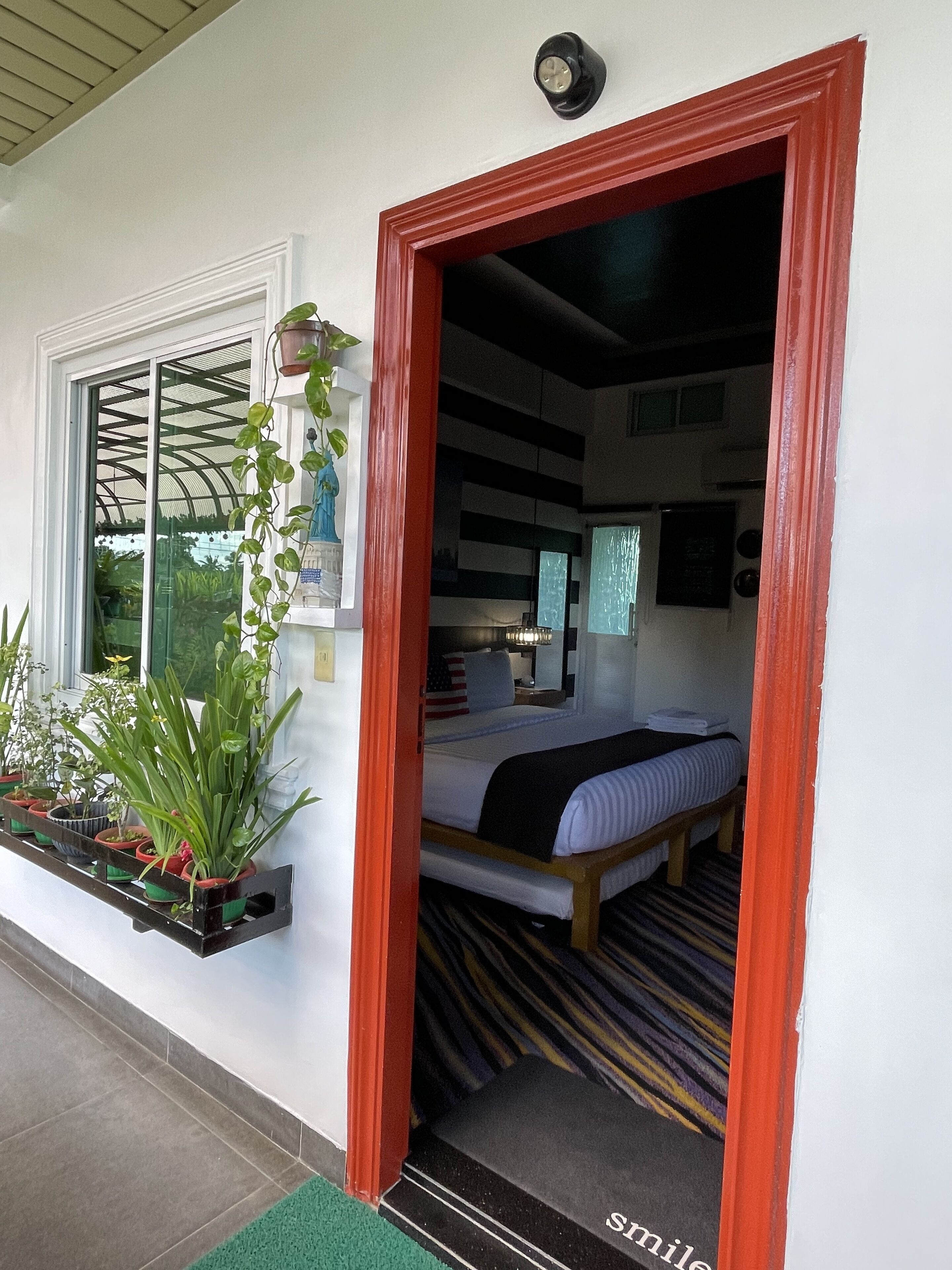 A room with a bed, a window with a view of greenery, and a red-framed doorway.
