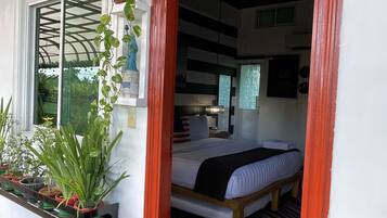A room with a bed, a window with a view of greenery, and a red-framed doorway.