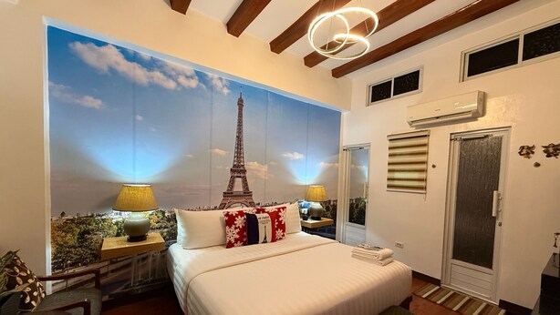 Een slaapkamer met een groot muurdecoratie van de Eiffeltoren, een bed met wit beddengoed, twee bedlampen, een houten bankje en een kleine tafel met een plant.