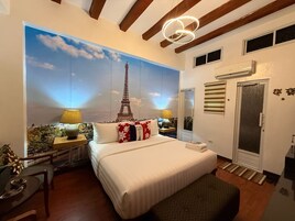 Un dormitorio con un gran mural de la Torre Eiffel, una cama con ropa de cama blanca, dos lámparas de noche, un banco de madera y una pequeña mesa con una planta.