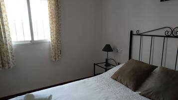 Una cama bien hecha con almohadas blancas y marrones, una mesita de noche con una lámpara y una ventana con cortinas de flores.