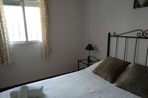 A neatly made bed with white and brown pillows, a small bedside table with a lamp, and a window with floral curtains. - Alojamiento Rural El Oregano (Campillos)