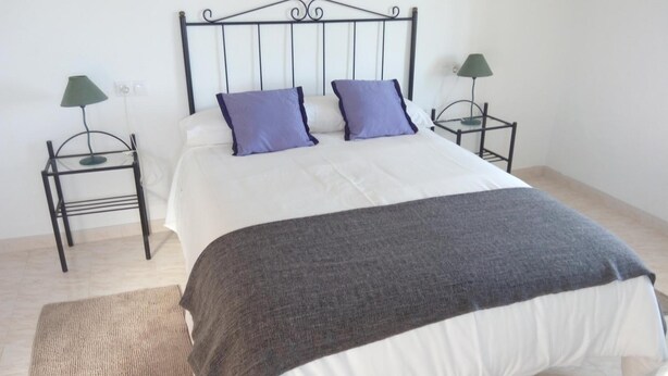 A bed with a metal headboard, two nightstands with lamps, a grey throw blanket, and a beige rug. - Alojamiento Rural El Oregano (Campillos)