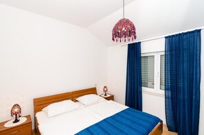 Un dormitorio con una cama de madera, ropa de cama blanca, una manta azul, una lámpara decorativa colgante y cortinas azules.