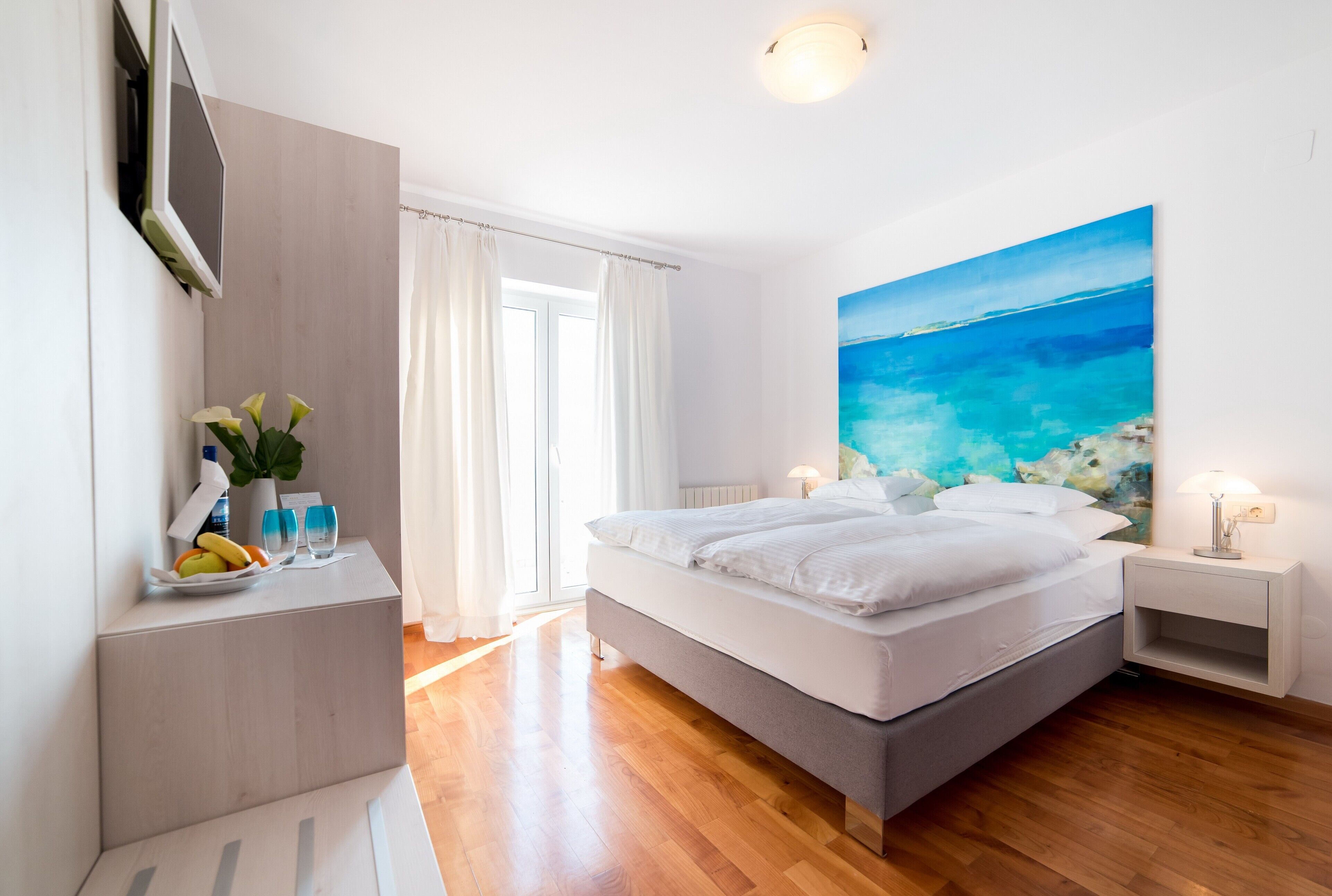 A modern bedroom with a large bed, a bedside table with a lamp, a painting of a beach scene, and a window with curtains.