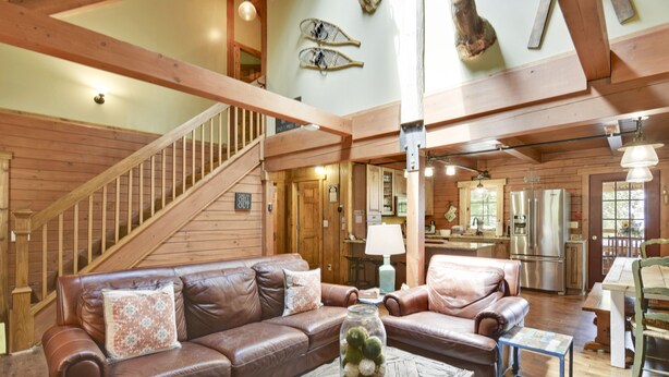 A spacious living area with a brown leather sofa, a coffee table with a glass jar, wooden beams, a staircase, and a kitchen area in the background. - Secluded Cabin, Comfortable Lux, Valley View ~ Canaan Valley & Seneca (Harman)