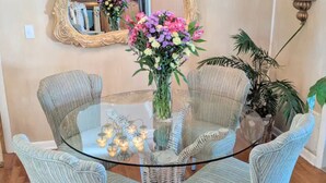Un comedor con una mesa redonda de vidrio rodeada por cuatro sillas de mimbre, un candelabro, un espejo y un jarrón de flores.