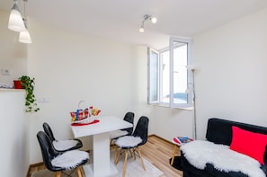 A modern, well-lit room with a dining table, two chairs, a black sofa with a red pillow, and a large window.