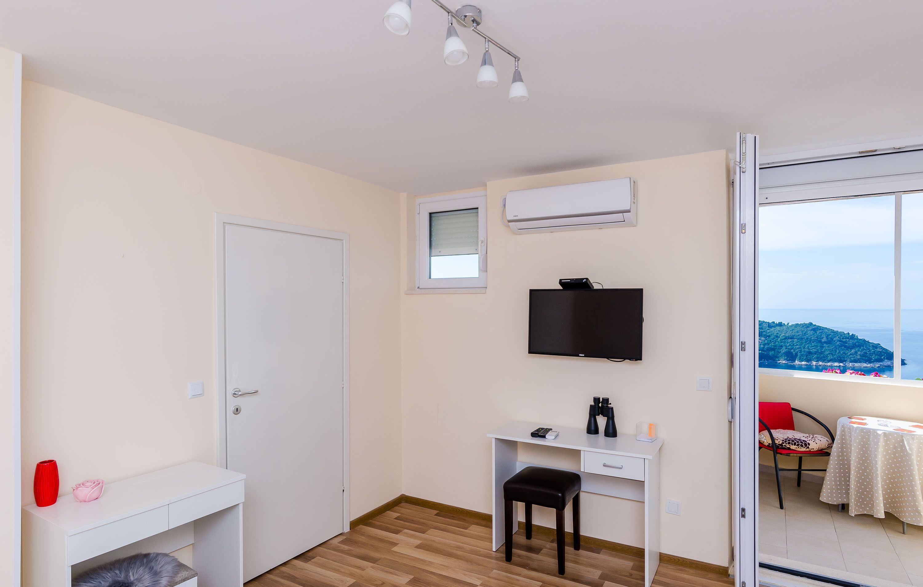 Una habitación pequeña y bien iluminada con un escritorio blanco, una silla, un televisor y un ventanal con vistas a un paisaje montañoso.