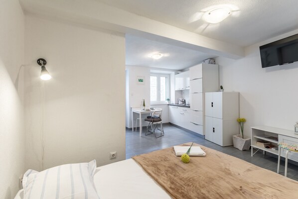 A modern hotel room with a bed, a wooden table, a desk, and a kitchenette.