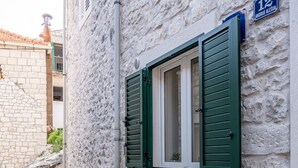 A narrow street with a white building featuring a window with green shutters, a small table, and two chairs. - Apartments Sonja - Studio (Pucisca)