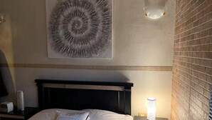 A neatly made bed with white linens and decorative pillows, a wall-mounted abstract artwork, a bedside table with a lamp, and a brick wall. - Very nice apartment F3 comfortable. (Agadir)