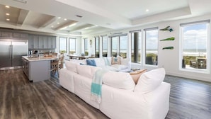 A spacious living area with a large white sofa, a kitchen with grey cabinets, and a view of the ocean through large windows.