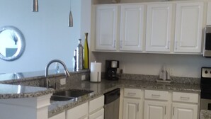 Una cocina con gabinetes blancos, encimera de granito, pileta de acero inoxidable, cafetera y lavavajillas.