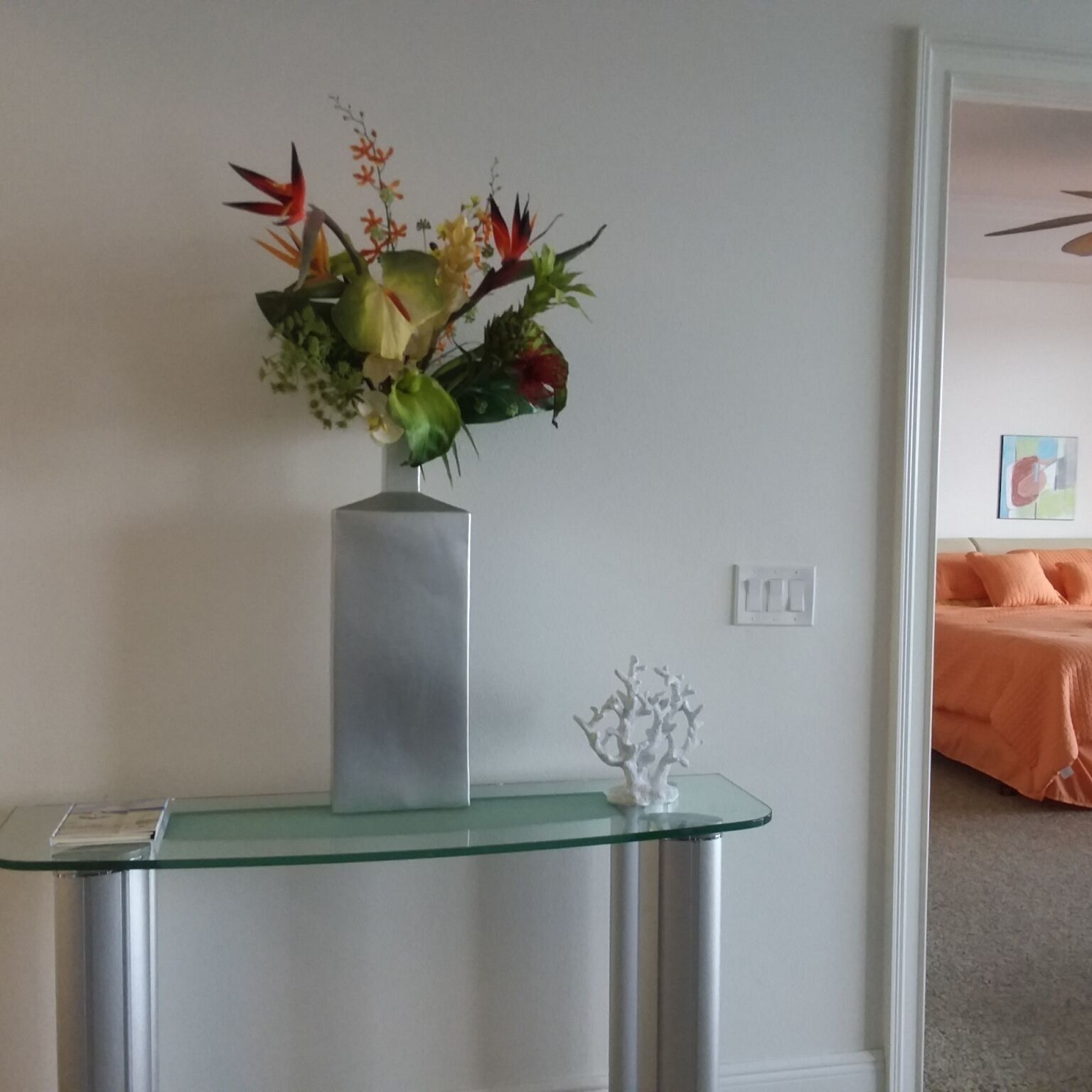 Una habitación con una mesa consola de vidrio que sostiene un florero con flores y una escultura blanca.