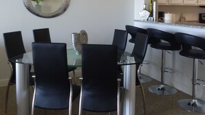 Un comedor con mesa de vidrio y sillas negras, lámpara colgante y área de cocina con armarios y encimera.