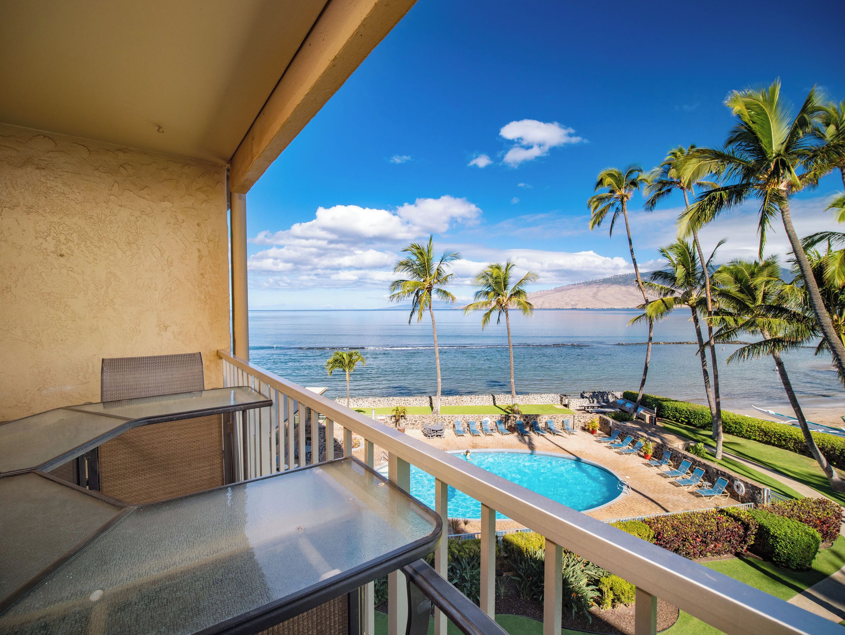 Ein Balkon mit Glastisch und Stühlen, Blick auf einen Poolbereich mit Liegestühlen und Palmen, mit Meerblick und einem entfernten Berg.