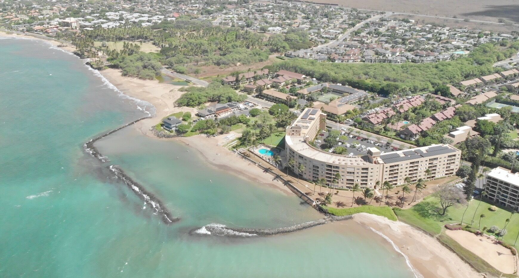 Vista aérea de um hotel à beira-mar, com uma extensa área de estacionamento e uma área residencial densa ao fundo.