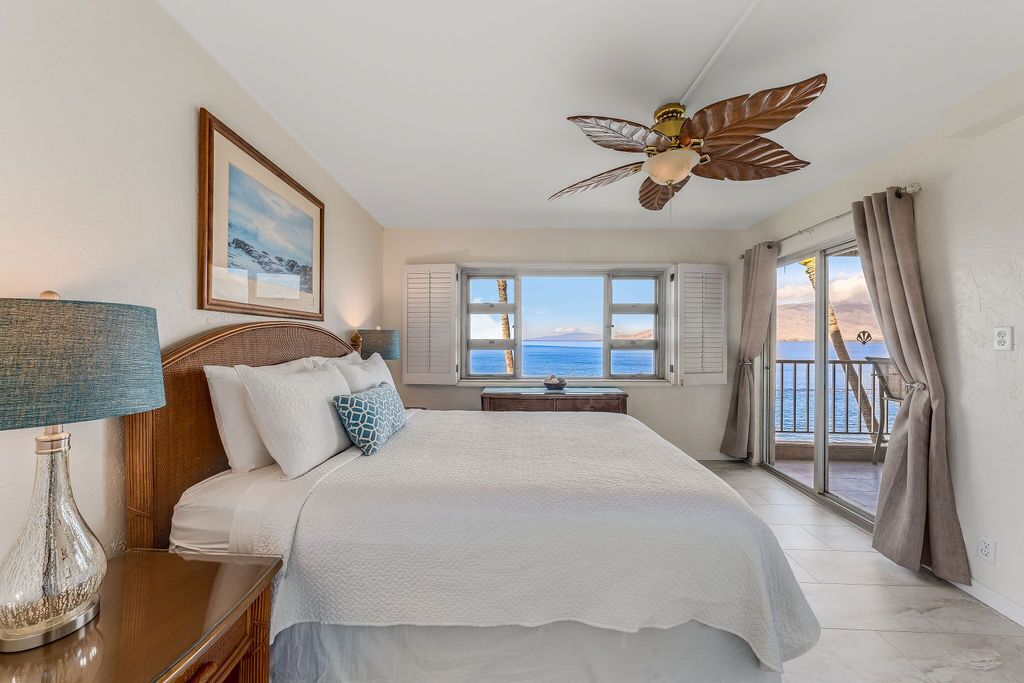 A bedroom with a large bed, a ceiling fan, a view of the sea through the window, and a balcony with curtains.