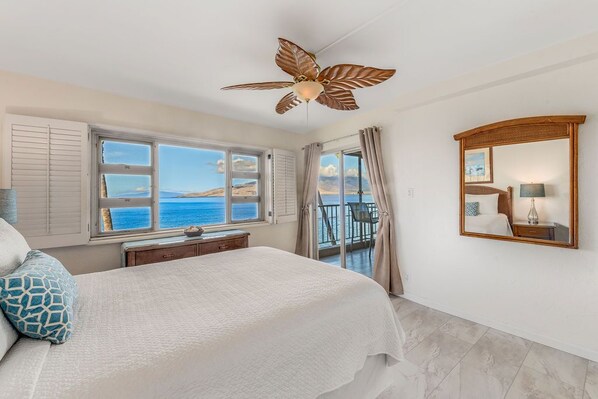 Un dormitorio con cama, ventilador de techo, ventana con vista al mar, balcón y espejo.