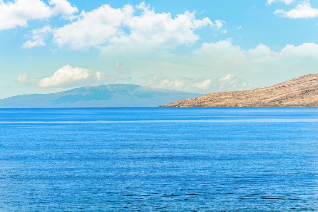 Paisaje costero con mar azul cristalino y montañas distantes bajo un cielo parcialmente nublado.