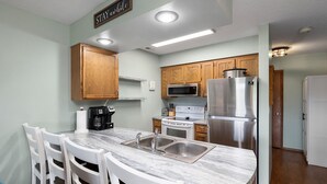 A compact kitchen with wooden cabinets, a stainless steel refrigerator, a microwave, a stove, and a coffee maker.