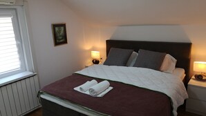 Una cama bien hecha con una colcha blanca y granate, dos almohadas y una toalla enrollada en el cabecero. Hay dos lámparas de noche, un cuadro en la pared y una ventana con persianas.